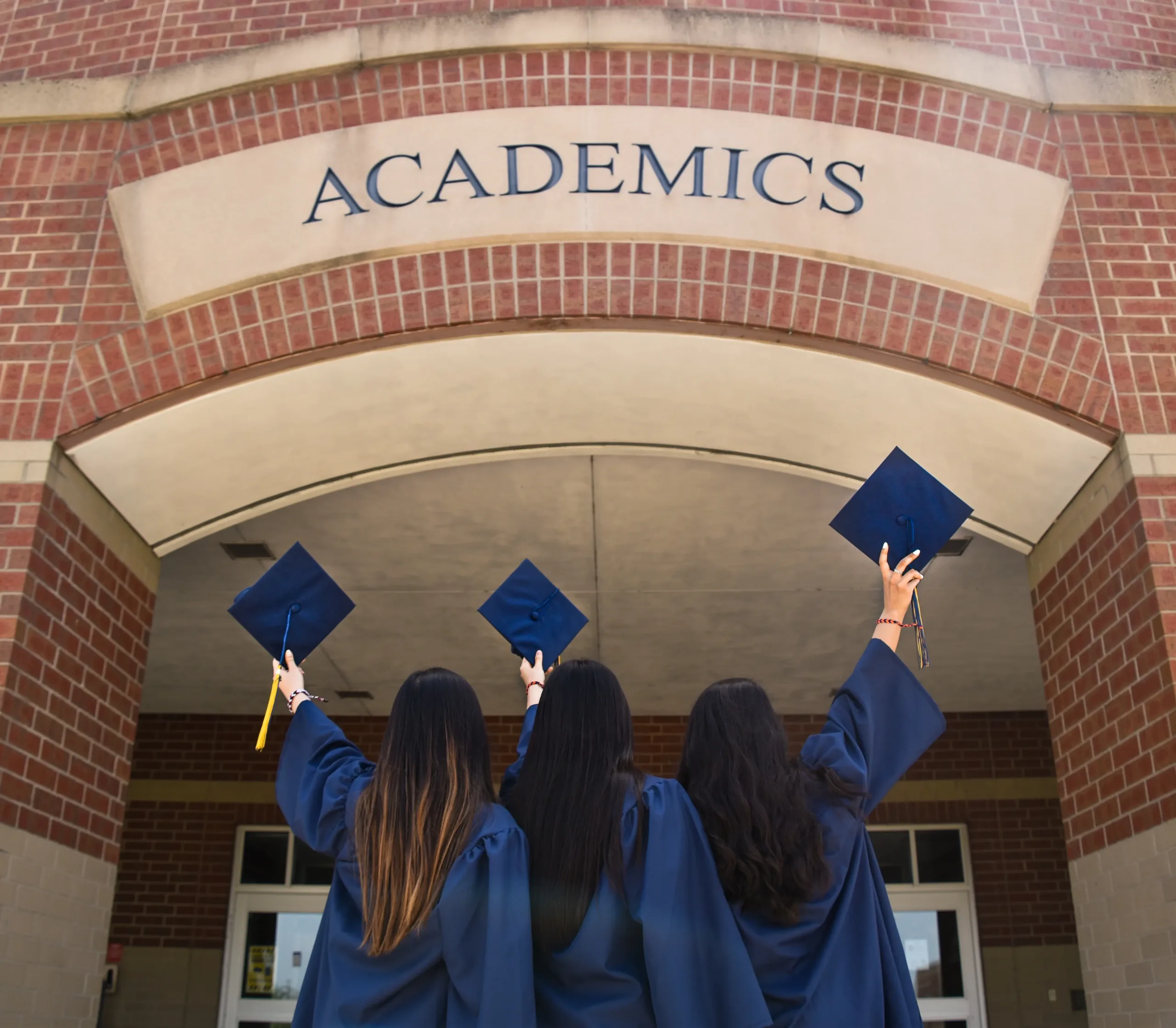 Graduates holding their caps in the air toward a brick academic building that says "academics" over the archway.
